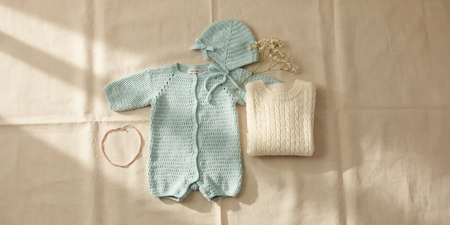 Handmade crochet baby set with a sage green romper and bonnet, cream cardigan sweater, and delicate crochet bracelet, flat lay for MapleHookCrochet